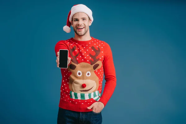 Handsome man in santa hat and christmas sweater showing smartphone with blank screen, isolated on blue — Stock Photo
