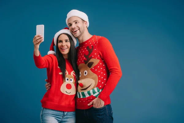 Alegre pareja en santa sombreros y navidad suéteres tomando selfie en smartphone aislado en azul - foto de stock