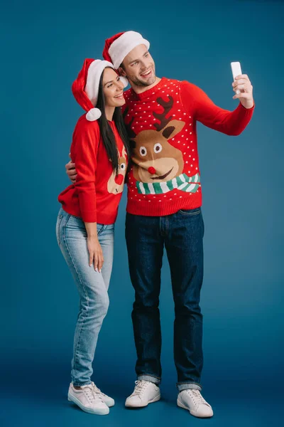 Feliz pareja en sombreros de santa y suéteres de Navidad tomando selfie en el teléfono inteligente aislado en azul - foto de stock