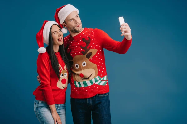Пара в шляпах Санта-Клауса и рождественских свитерах, торчащих языками, делая селфи на смартфоне, изолированном на голубом — стоковое фото