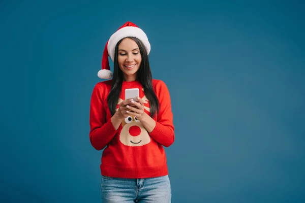 Jolie femme en chapeau de Père Noël et pull de Noël à l'aide d'un smartphone isolé sur bleu — Photo de stock