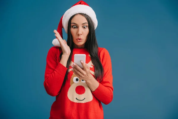 Attraktive überraschte Frau in Weihnachtsmütze und Weihnachtspullover mit Smartphone isoliert auf blau — Stockfoto