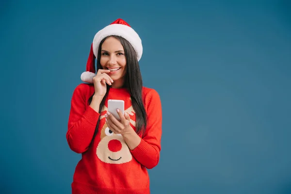 Lächelndes Mädchen mit Weihnachtsmütze und Weihnachtspullover mit Smartphone auf blauem Grund — Stockfoto