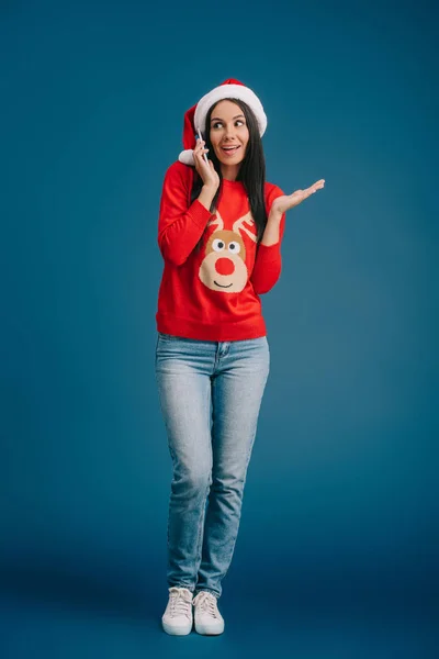 Happy woman in santa hat and christmas sweater talking on smartphone isolated on blue — Stock Photo