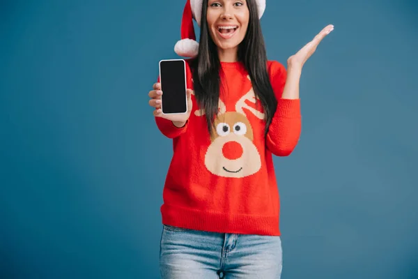 Mulher animado em chapéu de santa e camisola de Natal mostrando smartphone com tela em branco isolado em azul — Fotografia de Stock