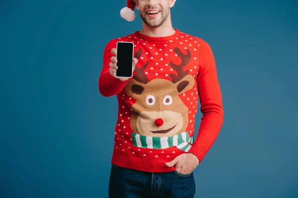 Vista recortada del hombre en sombrero de santa y suéter de Navidad que muestra el teléfono inteligente con pantalla en blanco, aislado en azul - foto de stock