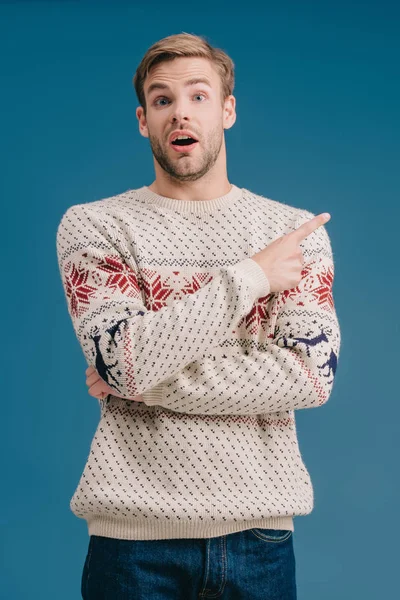 Handsome shocked man in winter sweater pointing at something isolated on blue — Stock Photo