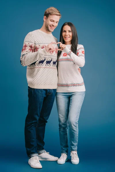 Casal sorridente em camisolas de inverno mostrando sinal de coração isolado em azul — Fotografia de Stock