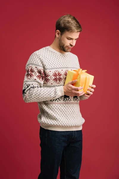 Guapo hombre reflexivo sosteniendo la caja de regalo de Navidad aislado en rojo - foto de stock