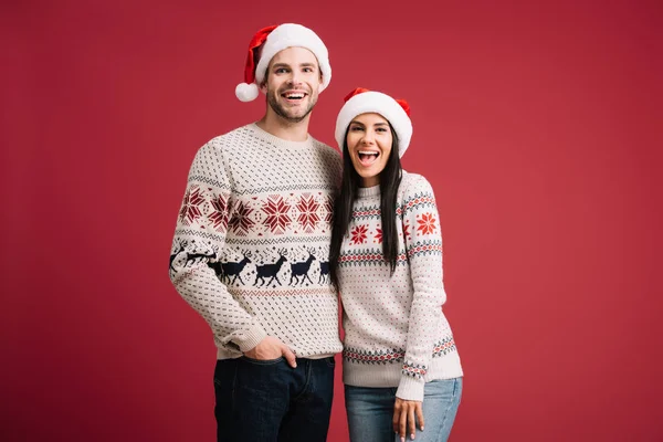 Belo casal animado posando em chapéus de santa e camisolas isoladas no vermelho — Fotografia de Stock