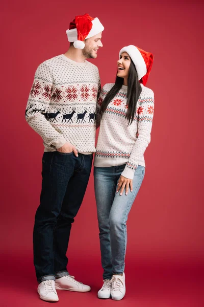 Felice coppia che abbraccia in cappelli di Babbo Natale e maglioni isolati su rosso — Foto stock