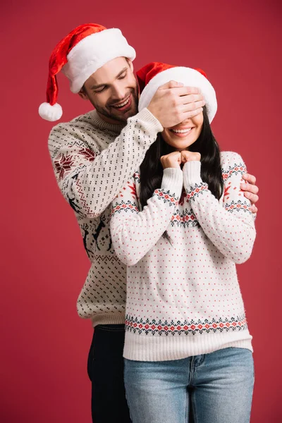 Smiling man in santa hat closing eyes of happy girlfriend to make surprise, isolated on red — Stock Photo