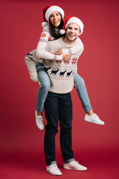 Glücklicher Freund huckepack Freundin in Weihnachtsmütze an Weihnachten auf rot — Stockfoto