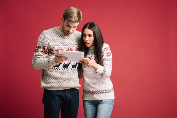 Pareja sorprendida en suéteres de invierno utilizando tableta digital aislada en rojo — Stock Photo