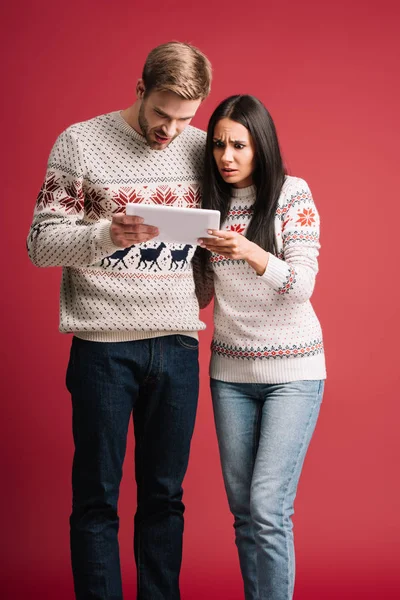 Shocked couple in winter sweaters using digital tablet isolated on red — Stock Photo