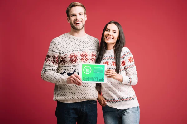 Улыбающаяся пара в зимних свитерах показывает цифровой планшет с приложением для покупок, изолированным на красном — стоковое фото