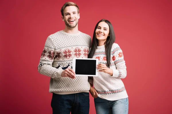 Coppia felice in maglioni invernali mostrando tablet digitale con schermo bianco isolato sul rosso — Foto stock