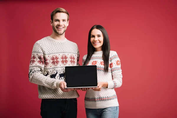 Красивая пара в зимних свитерах представляет ноутбук с чистым экраном, изолированным на красном — стоковое фото