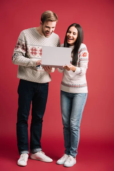 Beautiful laughing couple in winter sweaters using laptop isolated on red — Stock Photo