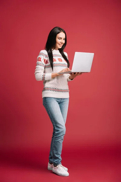 Donna sorridente in maglione invernale utilizzando computer portatile isolato su rosso — Foto stock