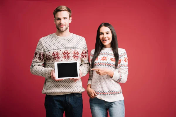 Paar in Winterpullovern zeigt auf digitales Tablet mit leerem Bildschirm auf rotem Hintergrund — Stockfoto