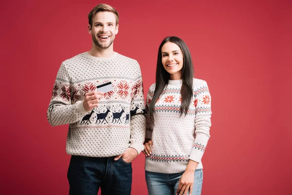 Couple in winter sweaters holding credit card for shopping isolated on red — Stock Photo