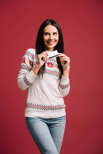 Beautiful smiling woman in winter sweater holding credit card, isolated on red — Stock Photo