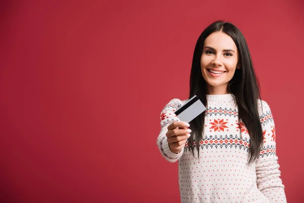 Cheerful woman in winter sweater holding credit card, isolated on red — Stock Photo