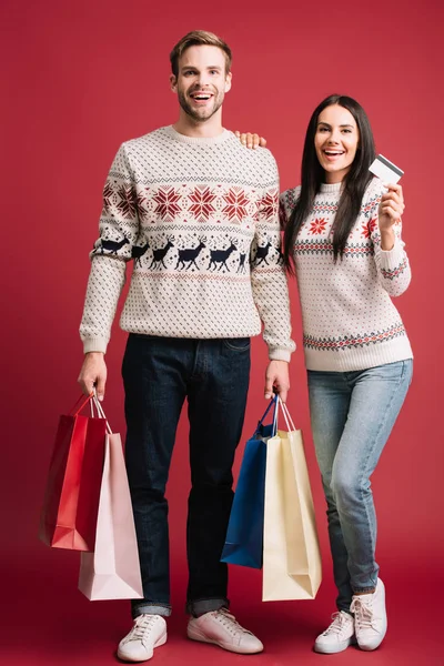 Casal sorridente em camisolas de inverno segurando cartão de crédito e sacos de compras isolados em vermelho — Fotografia de Stock