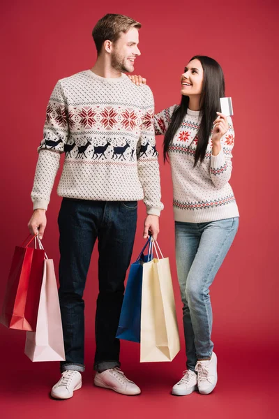 Namorada e namorado em camisolas de inverno segurando cartão de crédito e sacos de compras isolados no vermelho — Fotografia de Stock