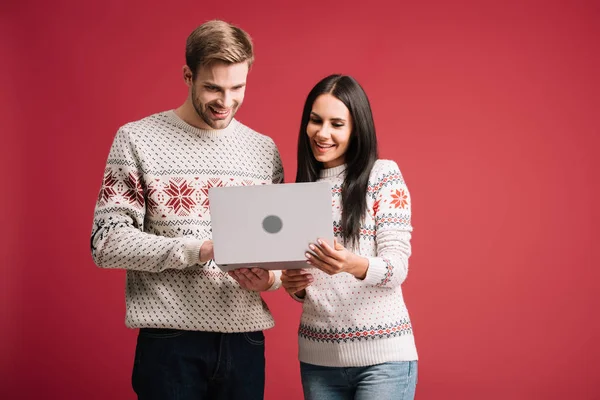 Красива усміхнена пара в зимових светрах використовує ноутбук ізольовано на червоному — стокове фото