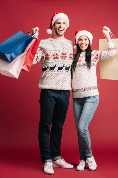 Coppia eccitata in cappelli di Babbo Natale tenendo borse della spesa isolate sul rosso — Foto stock