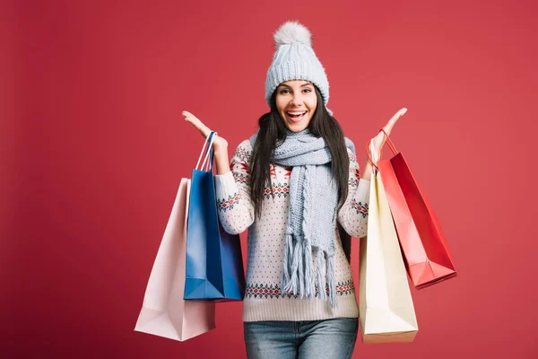 Mujer excitada en suéter de invierno, bufanda y sombrero sosteniendo bolsas de compras, aislado en rojo — Stock Photo