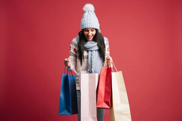 Femme heureuse en pull d'hiver, écharpe et chapeau regardant dans les sacs à provisions, isolé sur rouge — Photo de stock