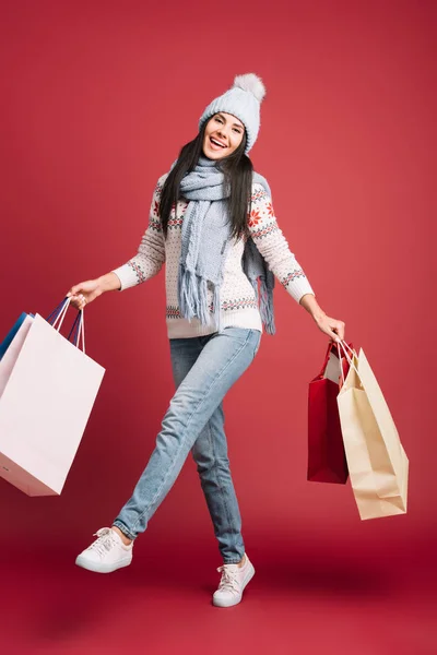 Bella ragazza in maglione invernale, sciarpa e cappello in possesso di borse della spesa, isolato su rosso — Foto stock