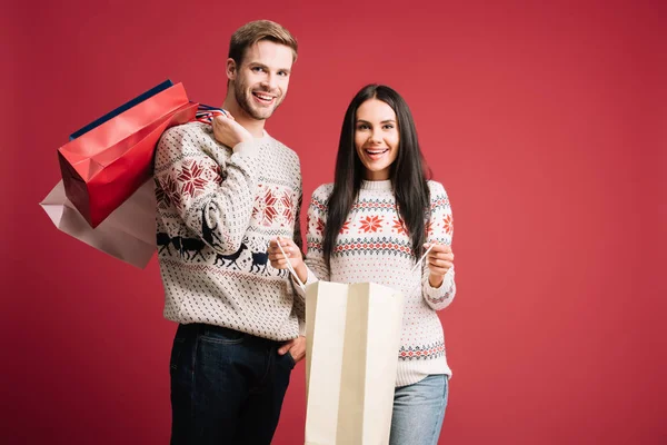 Щаслива пара в зимових светрах тримає сумки для покупок ізольовані на червоному — стокове фото