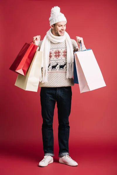 Fröhlicher Mann in Winterschal, Mütze und Pullover mit Einkaufstaschen, vereinzelt auf rotem Grund — Stockfoto