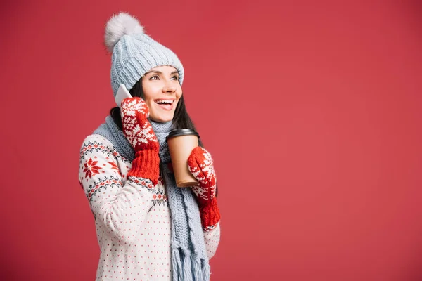 Приваблива жінка в зимовому одязі говорить на смартфоні і тримає каву, щоб піти ізольовано на червоному — стокове фото