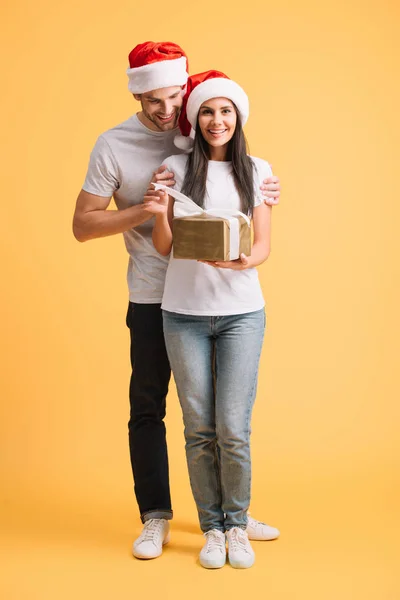 Couple joyeux chapeaux santa tenant Noël présent isolé sur jaune — Photo de stock