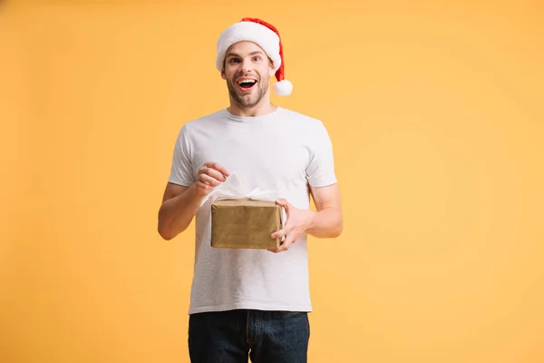 Збуджений чоловік у капелюсі Санта тримає подарунок Різдва ізольований на жовтому — стокове фото