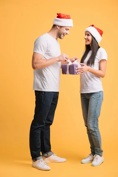 Paar in Weihnachtsmützen öffnet Weihnachtsgeschenk isoliert auf gelb — Stockfoto