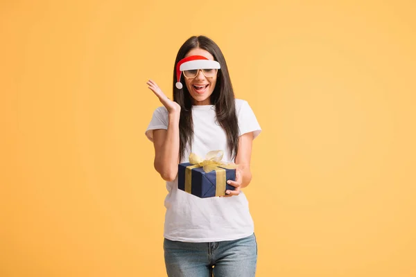 Überraschte Frau in Brille mit Weihnachtsmütze blickt auf Weihnachtsgeschenk isoliert auf gelb — Stockfoto