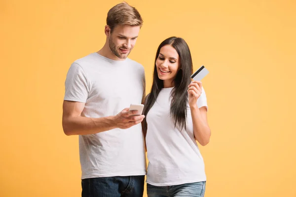 Beau couple faisant du shopping en ligne avec smartphone et carte de crédit isolé sur jaune — Photo de stock