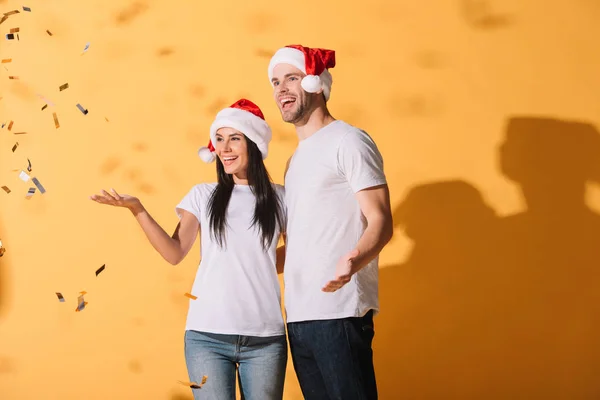 Pareja emocionada en sombreros de santa abrazándose en amarillo con confeti dorado - foto de stock