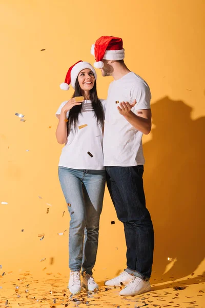 Schönes Paar in Weihnachtsmützen, die sich auf gelb mit goldenem Konfetti umarmen — Stockfoto