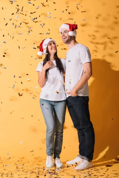 Fidanzato e fidanzata in cappelli di Babbo Natale abbracci su giallo con coriandoli dorati — Foto stock