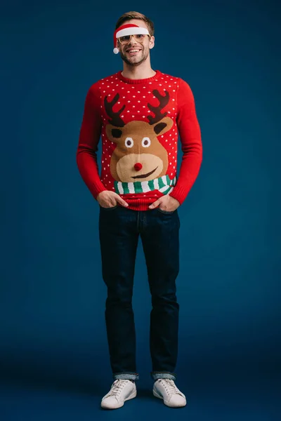 Happy man posing in christmas sweater and eyeglasses with santa hat, isolated on blue — Stock Photo