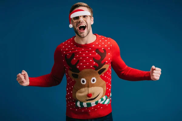 Взволнованный мужчина кричит и позирует в рождественском свитере и очках в шляпе Санты, изолированном на голубом — стоковое фото