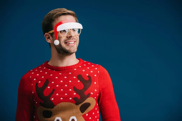 Lächelnder Mann posiert in Weihnachtspullover und Brille mit Weihnachtsmütze, isoliert auf blauem Grund — Stockfoto
