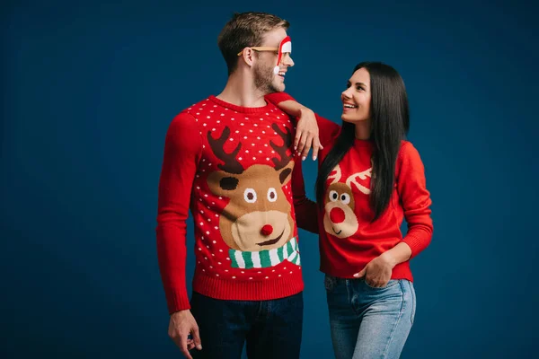 Smiling man in glasses with santa hat hugging girlfriend isolated on blue — Stock Photo